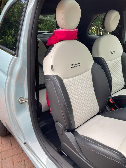 This image shows the interior of a Fiat 500 car, and a Hot Pink scarffio® stored in standby mode - tied around the headrest of the drivers seat, like a scarf. There is a small portion of the Hot pink scarffio® showing between the headrest and the top of the seat, and the rest is draped over the back of the seat, out of shot, except for a tiny bit peeping out from behind the bottom of the seat. 