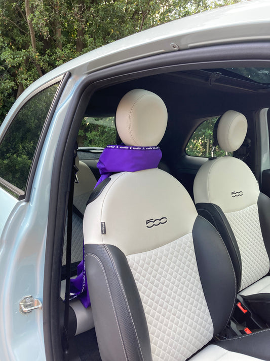 This image shows the interior of a Fiat 500 car, and a Rich Purple scarffio® stored in standby mode - tied around the headrest of the drivers seat, like a scarf. There is a small portion of the Rich Purple scarffio® showing between the headrest and the top of the seat, and the rest is draped over the back of the seat, out of shot, except for a tiny bit peeping out from behind the bottom of the seat. 