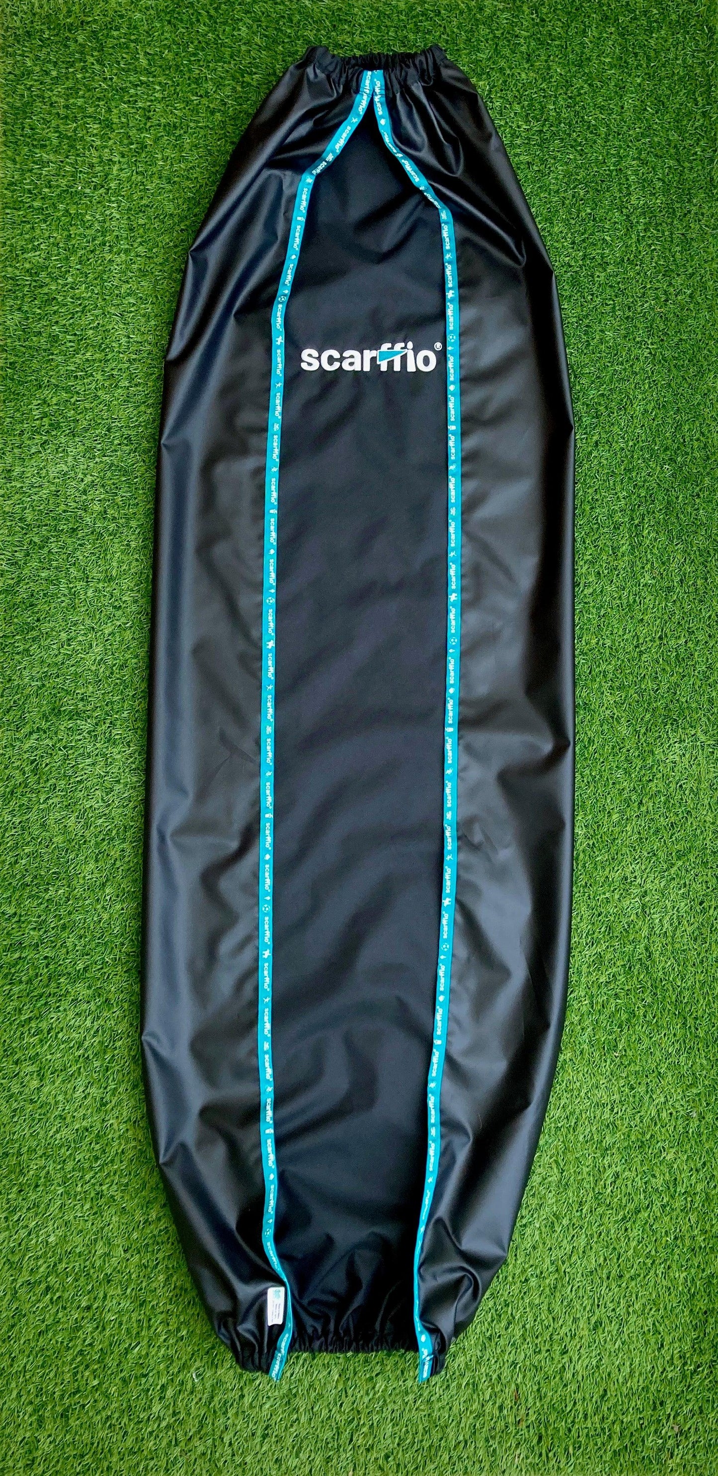 Shows a folded, full size  Custom Jet Black and Wild Teal  scarffio® on a grass background. The folding demonstrates the waterproof coating on the back of the scarffio® and the soft feel fabric on the front . Also shown is  the contrasting Wild Teal embroidered flag and branded edging strip.