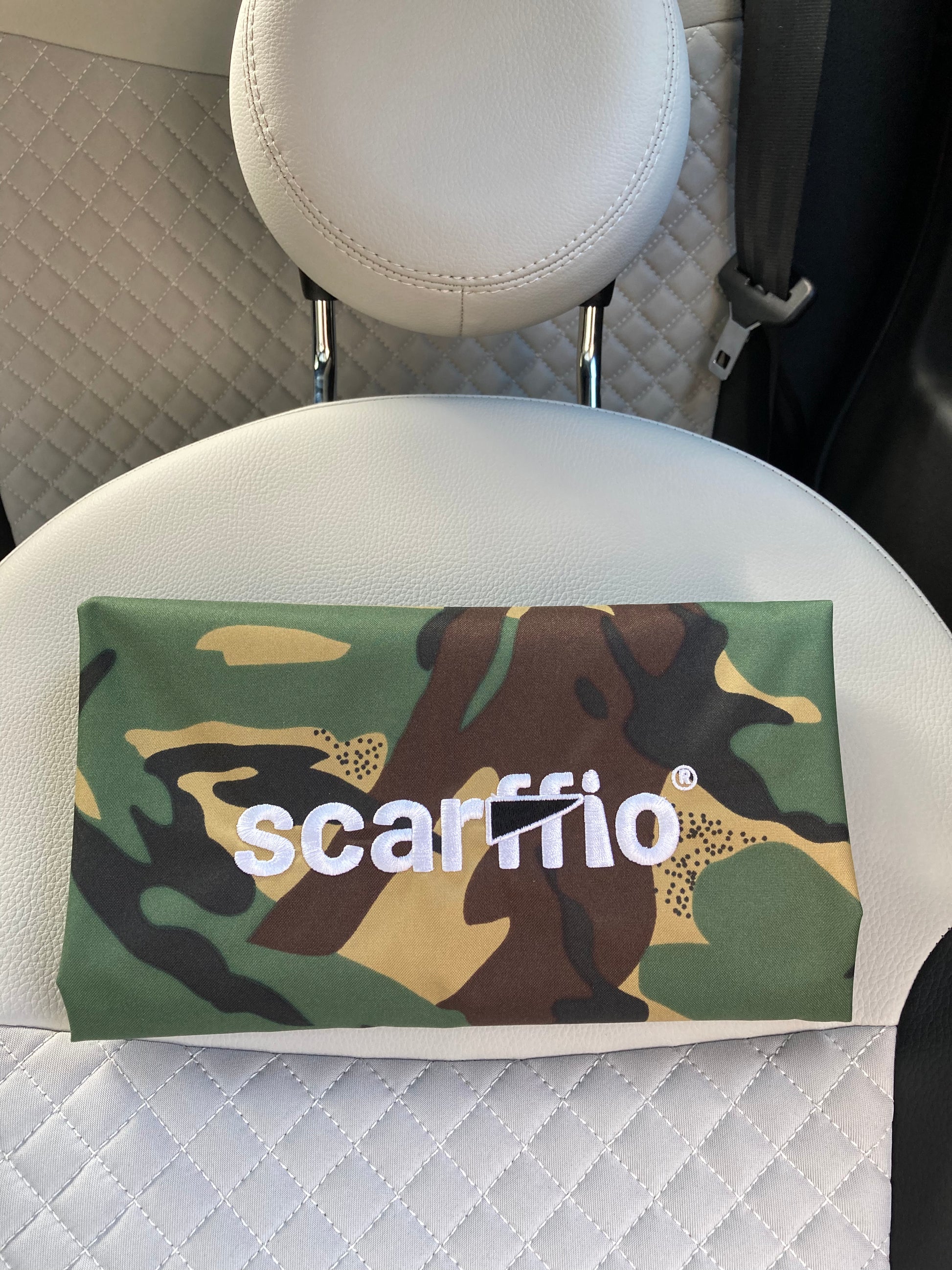 This image shows a a folded Camo scarffio®, resting on a cream and black quilted car seat. The focus of the image is  the white embroidered scarffio®  logo with its' contrasting black cape detail. 