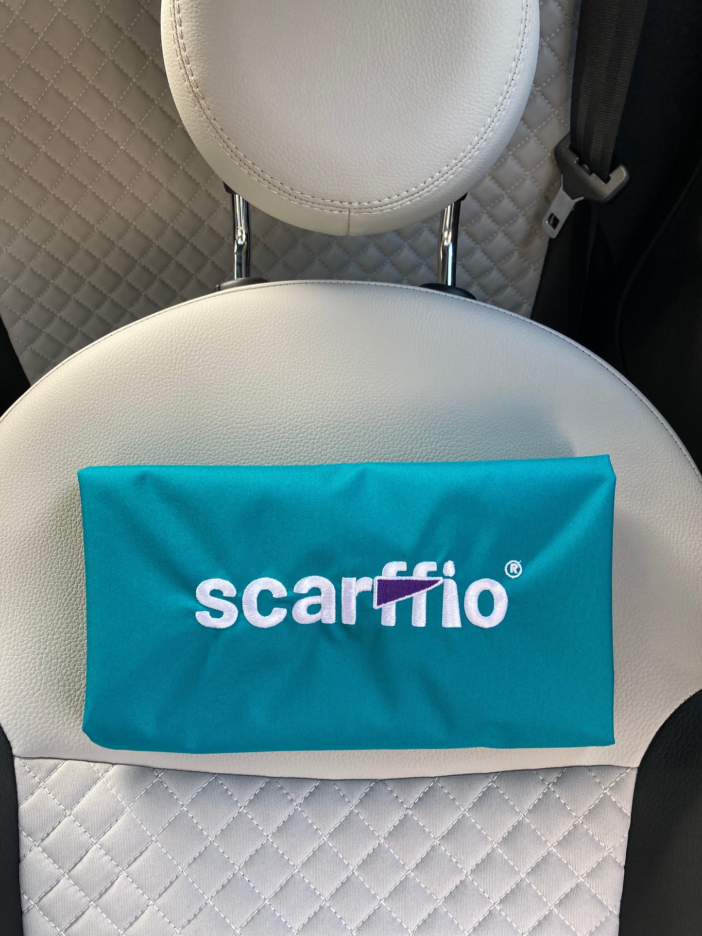 This image shows a a folded Hero scarffio®, resting on a cream and black quilted car seat. The focus of the image is  the white embroidered scarffio®  logo with its contrasting purple cape detail. 