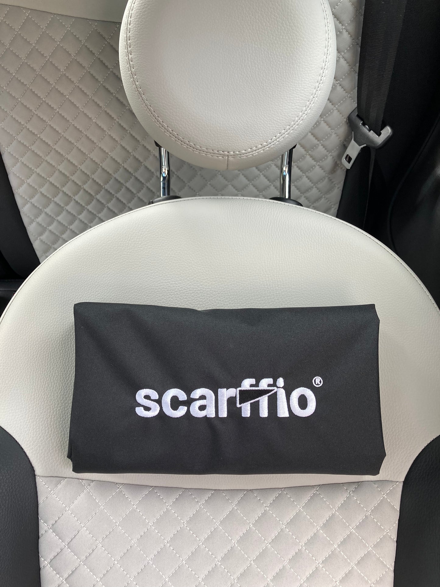 This image shows a a folded Jet Black scarffio®, resting on a cream and black quilted car seat. The focus of the image is  the white embroidered scarffio®  logo with its matching black cape detail. 