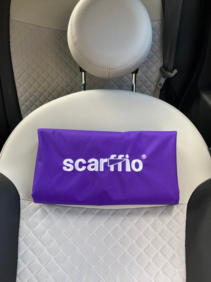This image shows a a folded Rich Purple scarffio®, resting on a cream and black quilted car seat. The focus of the image is  the white embroidered scarffio®  logo with its' matching purple cape detail. 