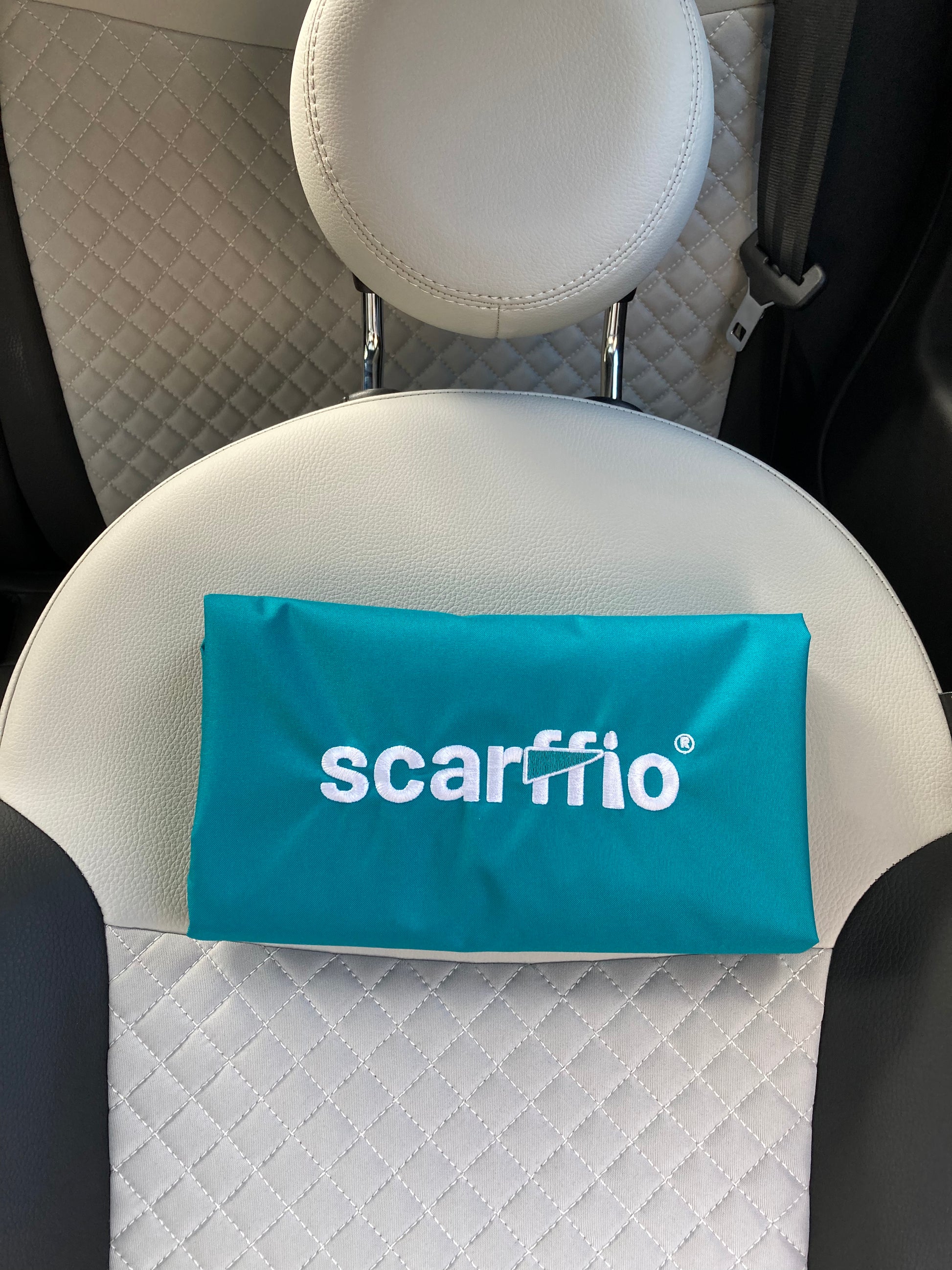 This image shows a a folded Wild Teal scarffio®, resting on a cream and black quilted car seat. The focus of the image is  the white embroidered scarffio®  logo with its' matching Teal cape detail. 