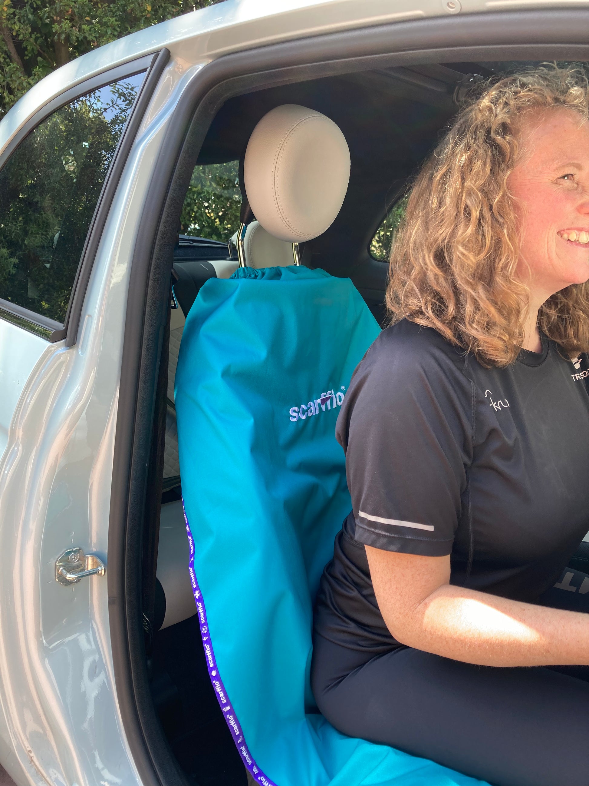 This image shows at the interior of a Fiat 500, with a Hero scarffio® down on the drivers seat , with a smiling female driver in situ. The Hero scarffio® has a Wild Teal base fabric with contrasting purple edging , and a contrasting purple cape on the embroidered central logo.  
