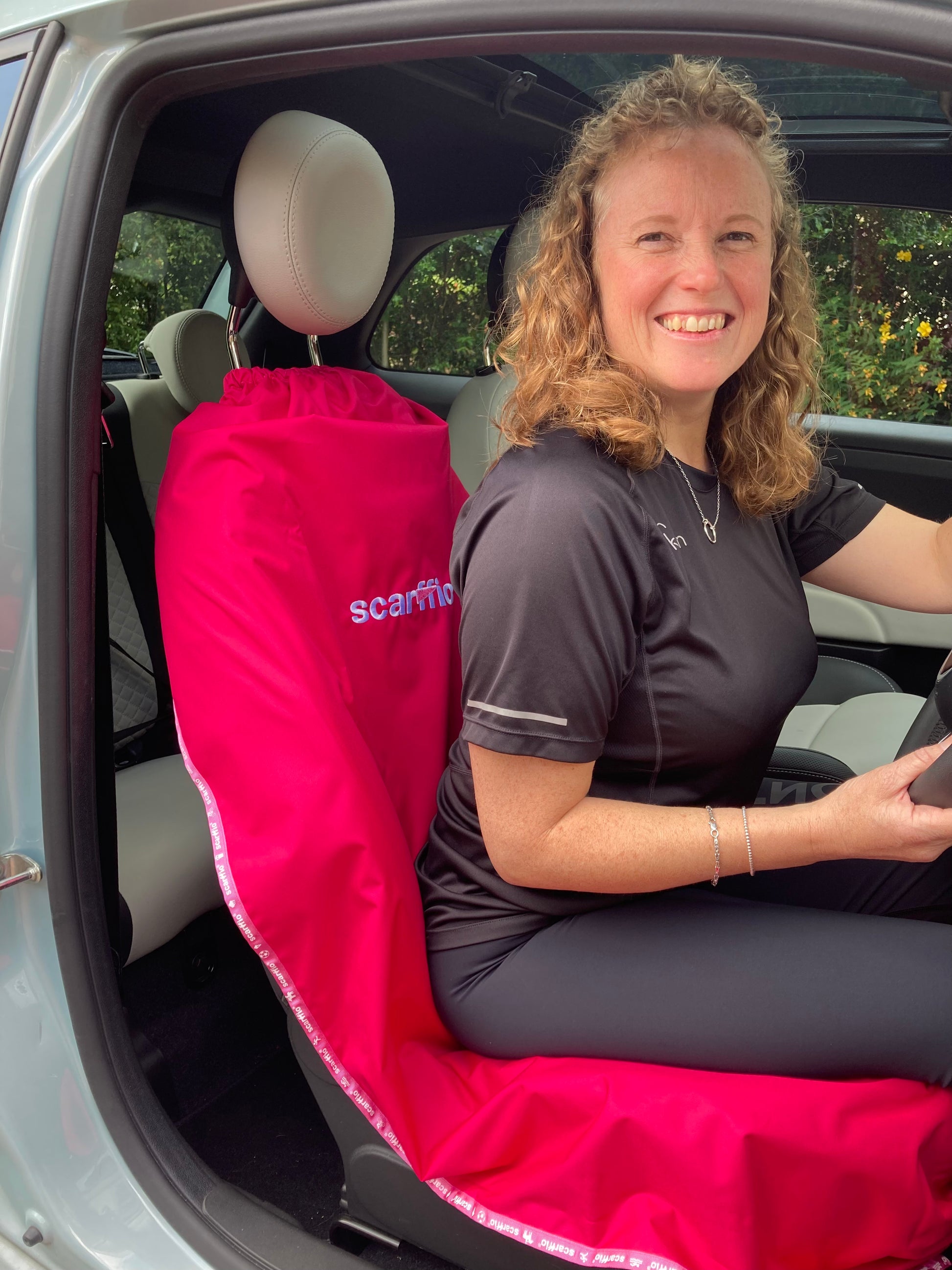This image shows at the interior of a Fiat 500, with a Hot Pink scarffio® down on the drivers seat , with a smiling female driver in situ. 